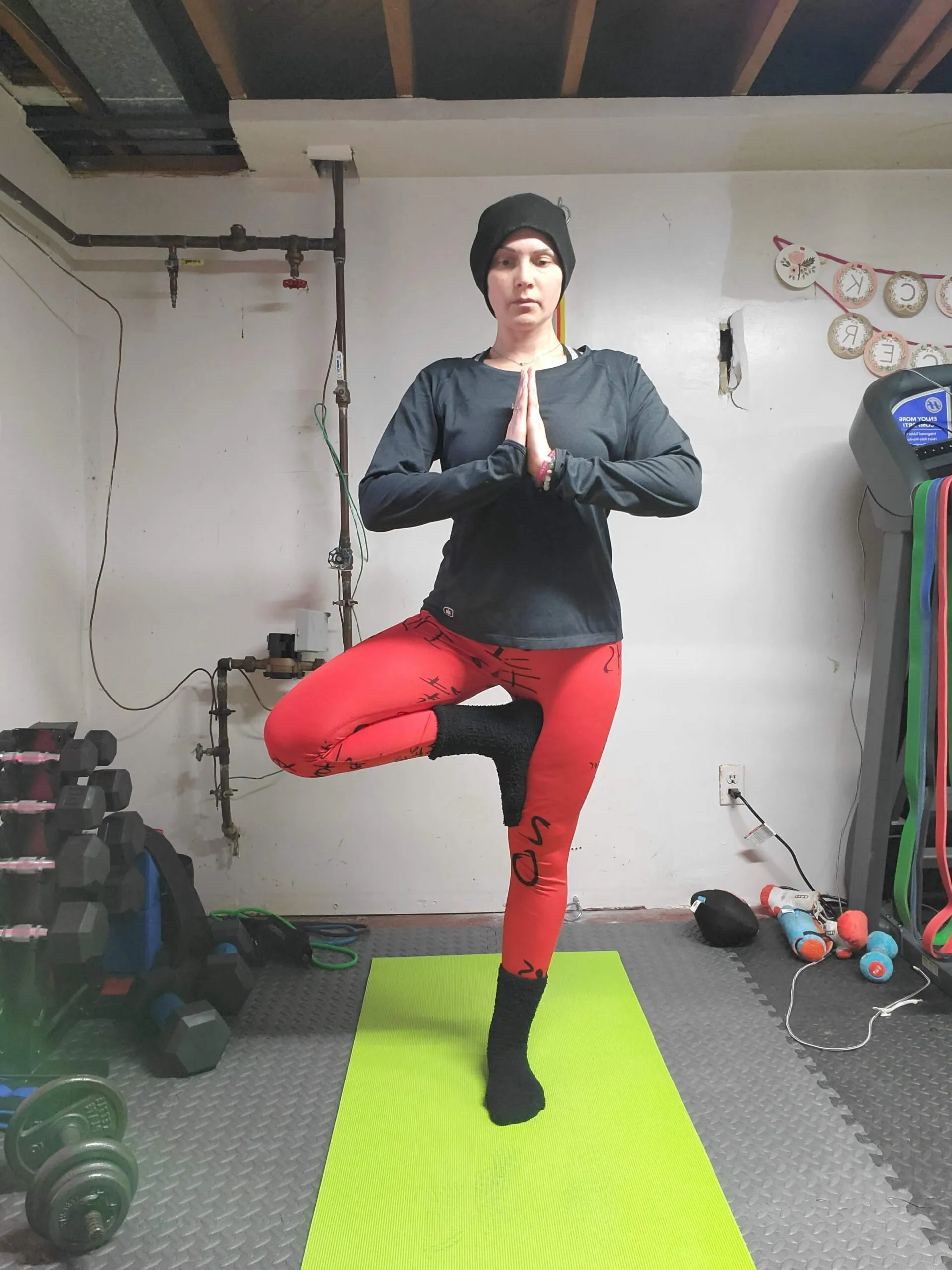 A woman in a gym standing on a yoga mat doing a yoga pose with praying hands and her right leg folded into her left leg.