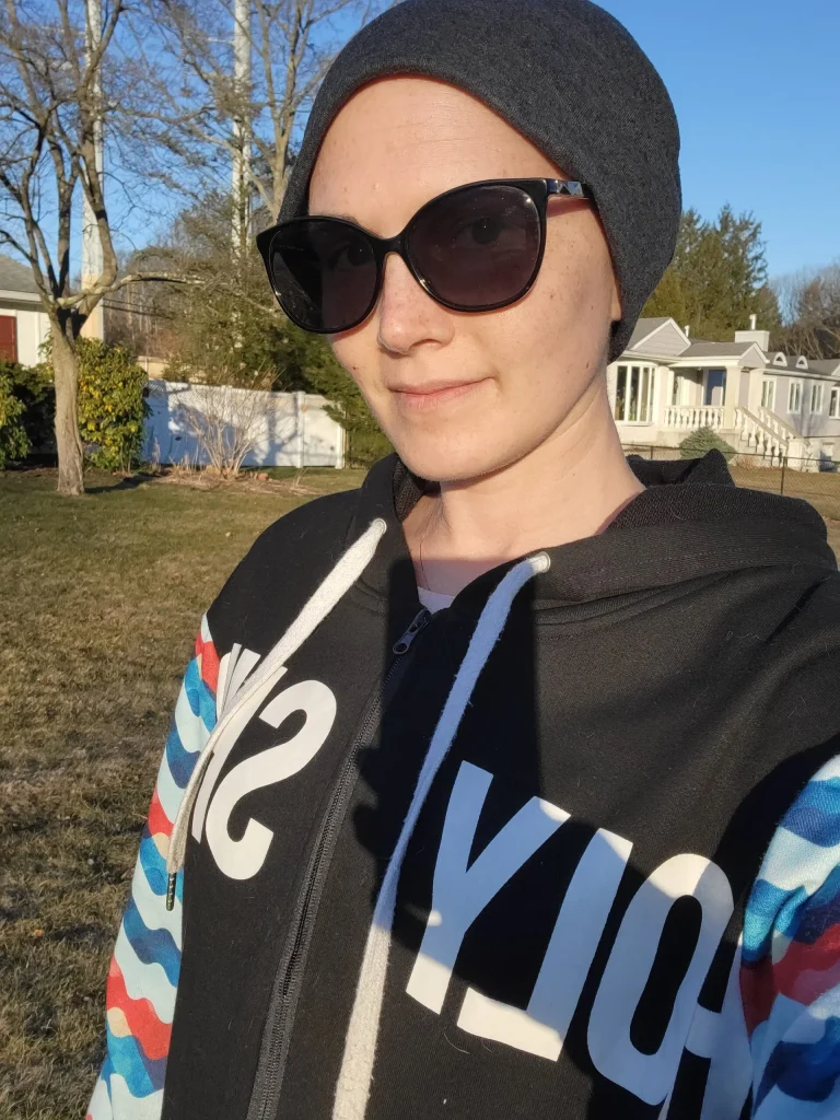A woman taking a selfie standing outside wearing a beanie, sunglasses and zip up sweatshirt.