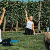A group of women outside sitting on yoga mats in a field with their right arm stretched up in the air.