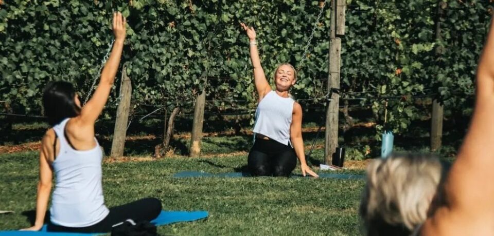 A group of women outside sitting on yoga mats in a field with their right arm stretched up in the air.