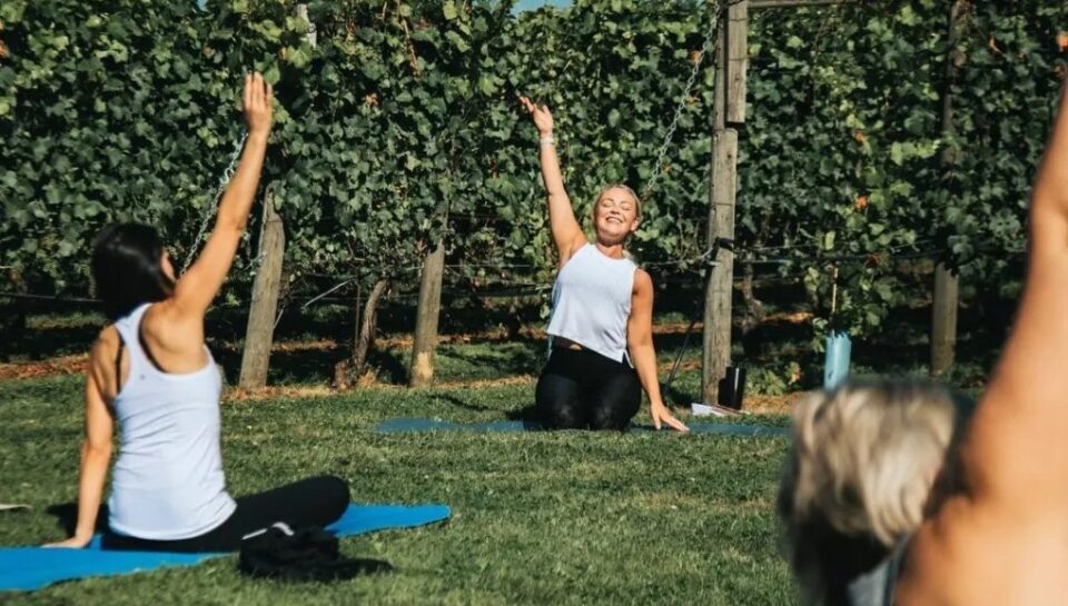 A group of women outside sitting on yoga mats in a field with their right arm stretched up in the air.
